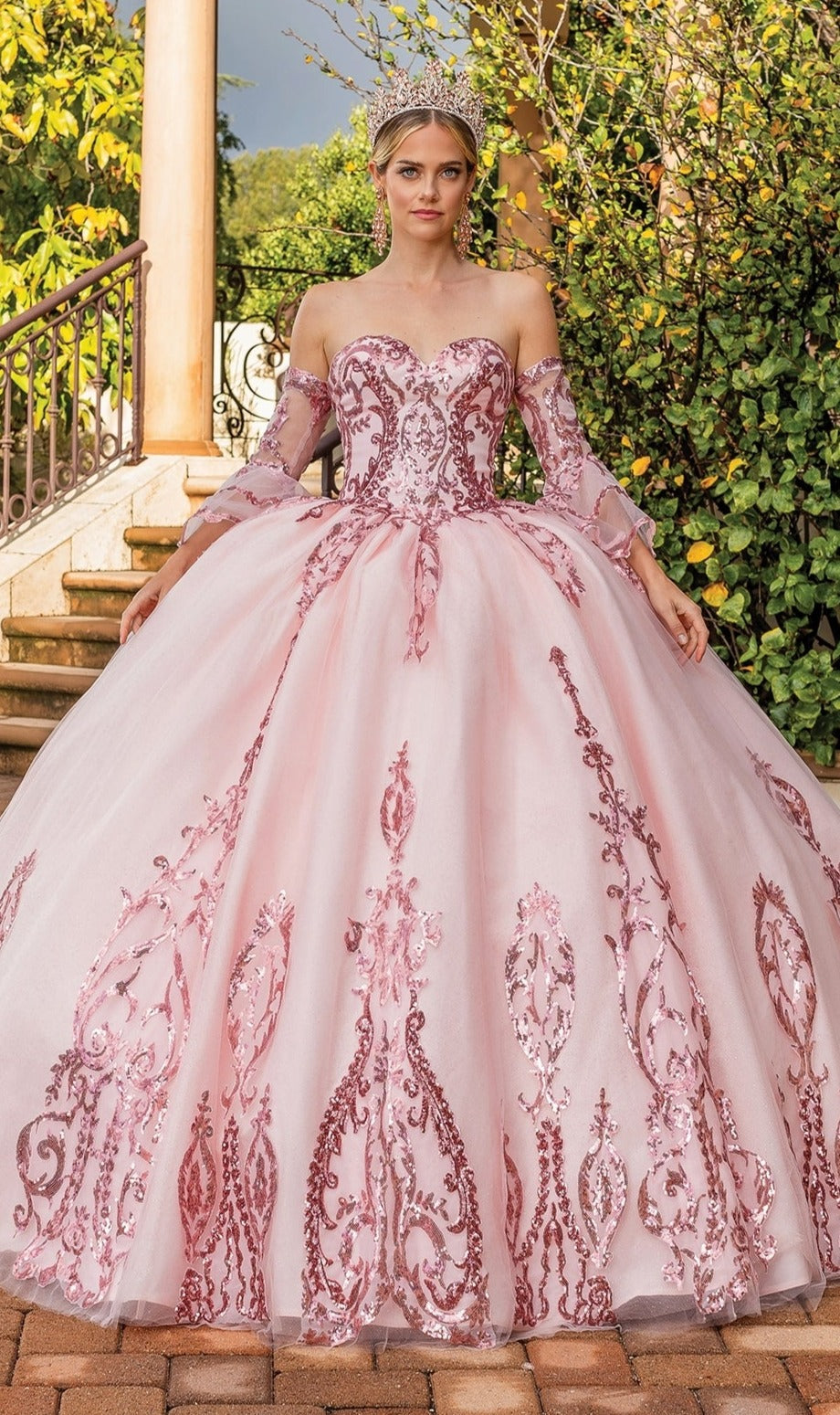 A woman wearing the Quinceanera Dress 1861 By Dancing Queen, a blush pink off-the-shoulder gown with intricate embroidery and a tiara, stands outside near steps.